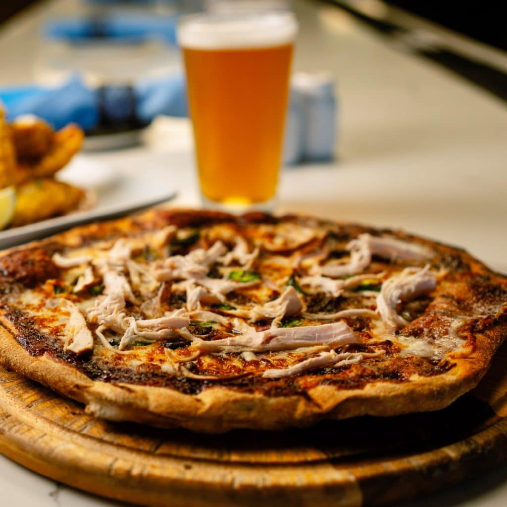 Pizza on a wooden board topped with shredded chicken and herbs, with a glass of beer blurred in the background.